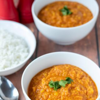 Two bowls of yellow lentil dahl one in front of the other garnished with coriander. A bowl of rice and spoons to the left hand side.