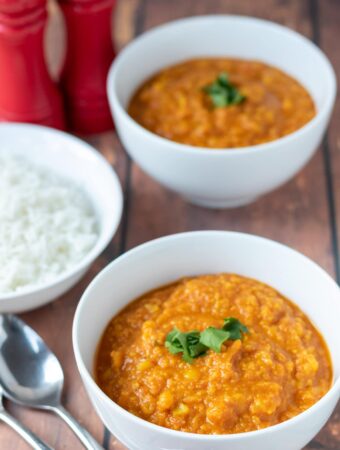 Two bowls of yellow lentil dahl one in front of the other garnished with coriander. A bowl of rice and spoons to the left hand side.