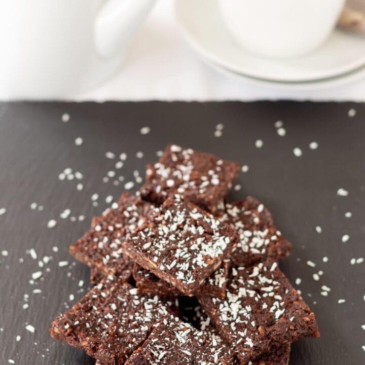 A stack of 5-ingredient no bake chocolate brownies on a slate decorated with grated coconut. A tea pot and tea cup at top of picture.
