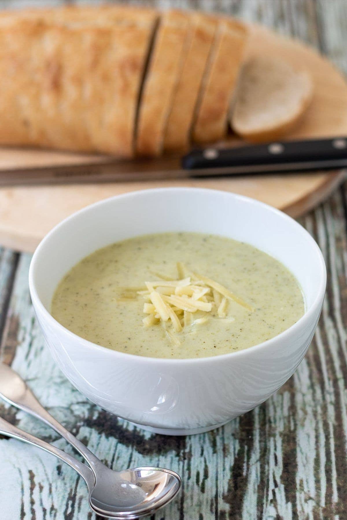 A bowl of broccoli and potato soup garnished with grated cheese and a spoon beside. Sliced bread on a board at the back.