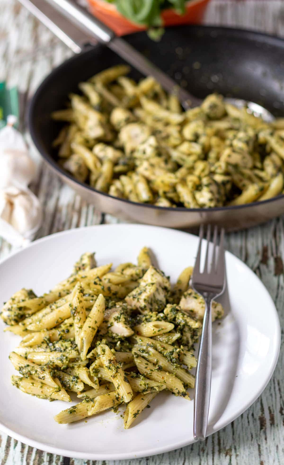 A plate of cooked chicken pesto pasta with a fork to the right hand side. A frying pan of the rest of the cooked dinner in the background.