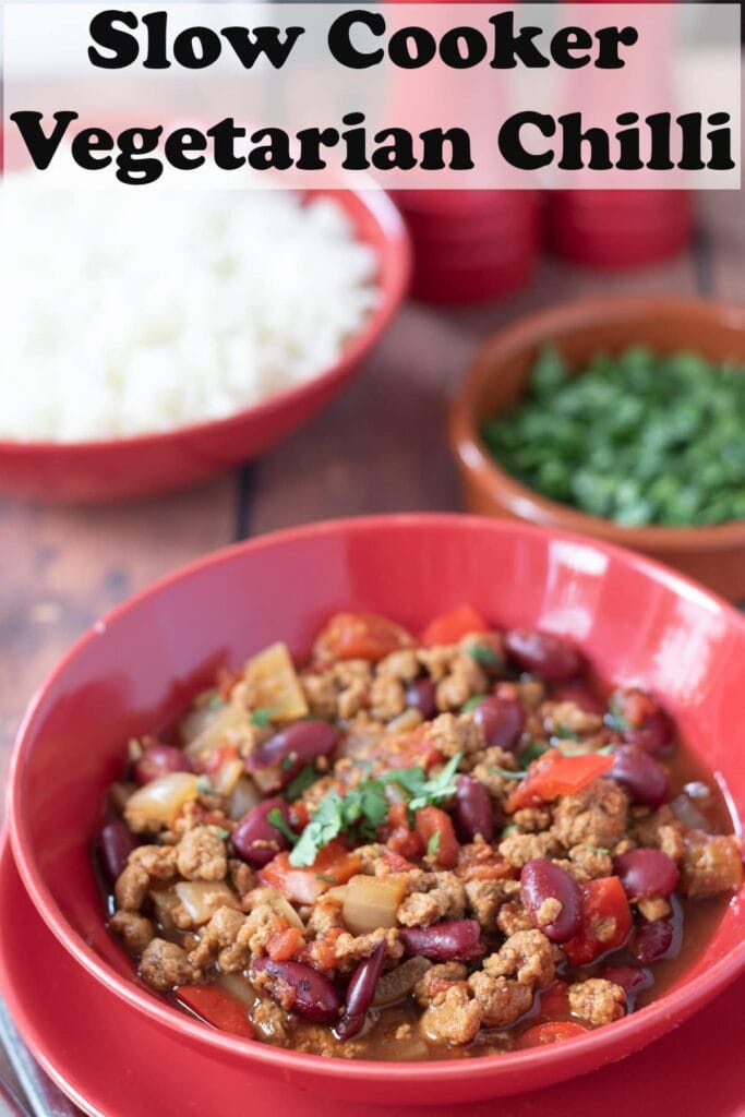 A bowl of slow cooker vegetarian chilli garnished with chopped coriander. A dish of chopped coriander and bowl of basmati rice in the background. Pin title text overlay at top.