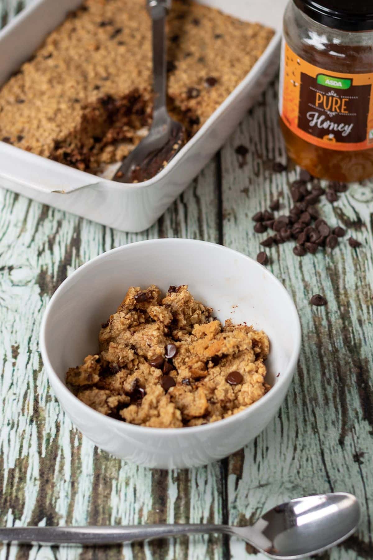 A bowls of chocolate peanut butter baked oats with a spoon in front. Rest of the recipe ins a casserole dish in the background with a jar of honey beside.
