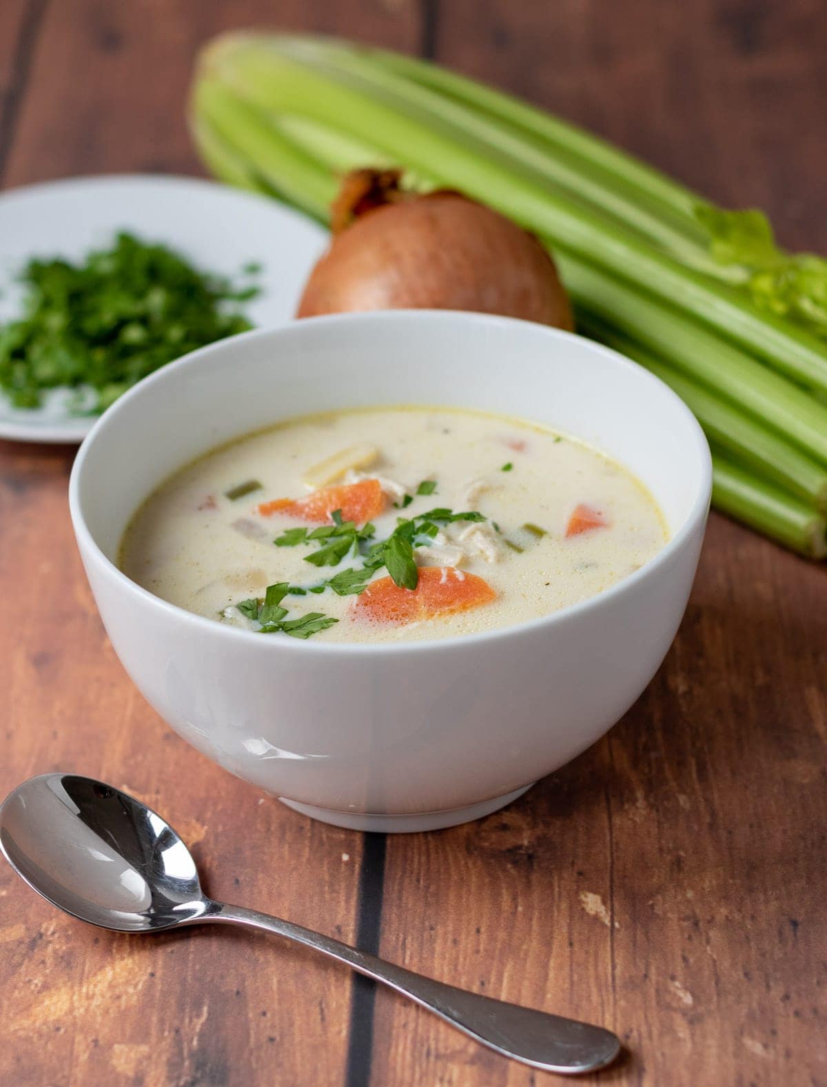 A bowl of healthy creamy chicken potato soup. Soup spoon in front, parsely, an onion and bunch of celery in the background.