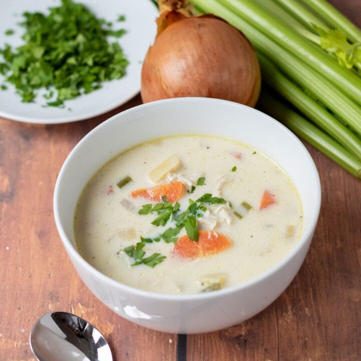 A bowl of healthy creamy chicken potato soup with a soup spoon in front and an onion, bunch of celery and plate of chopped parsley in the background as decoration.