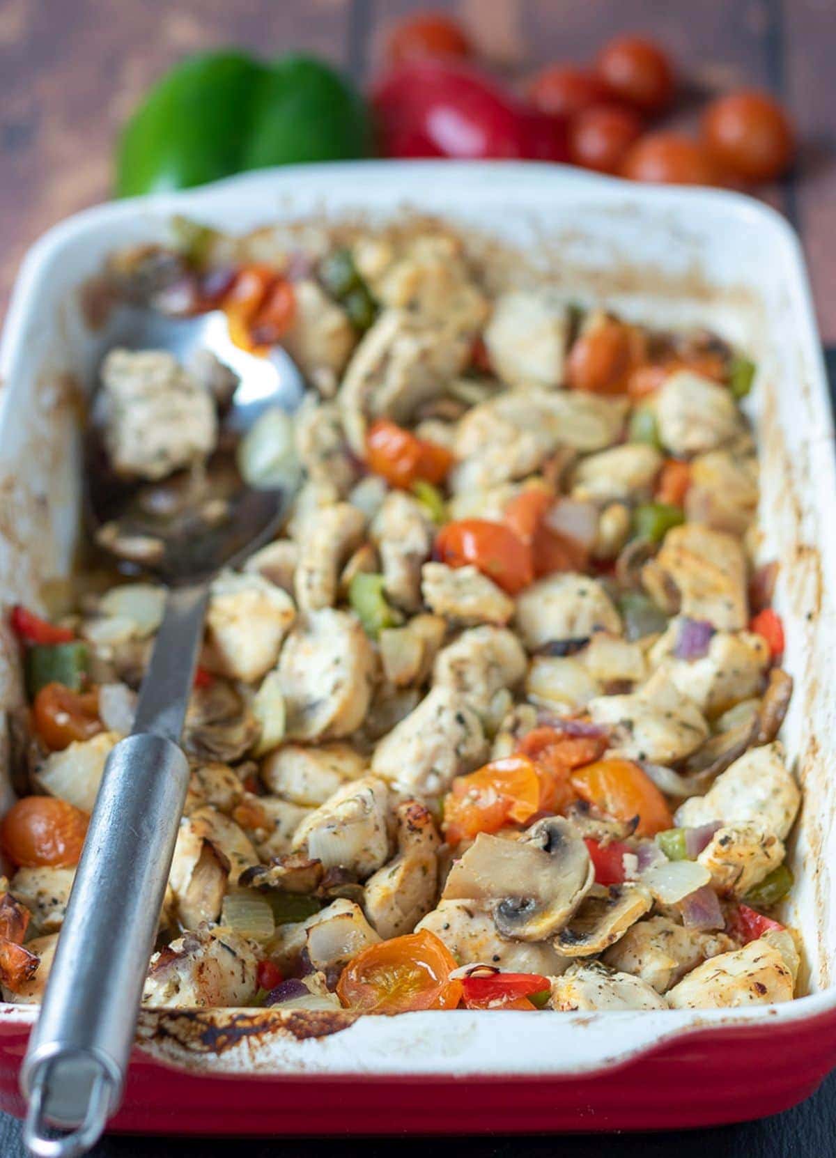 Baked chicken and mixed vegetables cooked in a stoneware serving dish with a serving spoon to the left hand side.