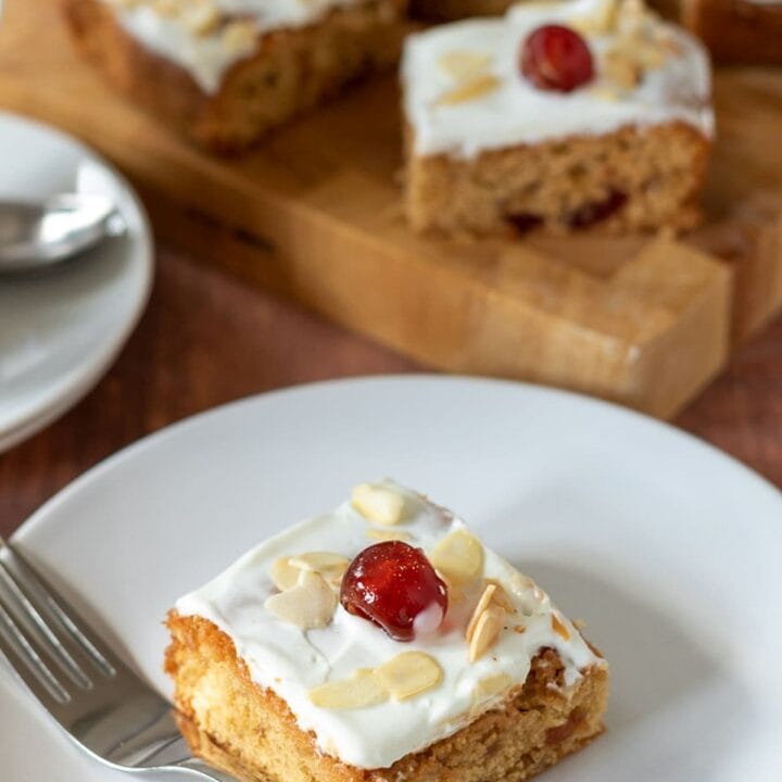 A square of cherry bakewell tray bake served on a white plate with a fork to the side. Chopping board with the rest of the tray bake squares on in the background.