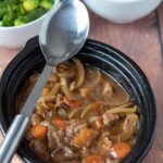 Slow cooker beef and ale stew with a serving spoon placed over the top. Side dishes of broccoli and mashed potatoes at the top.