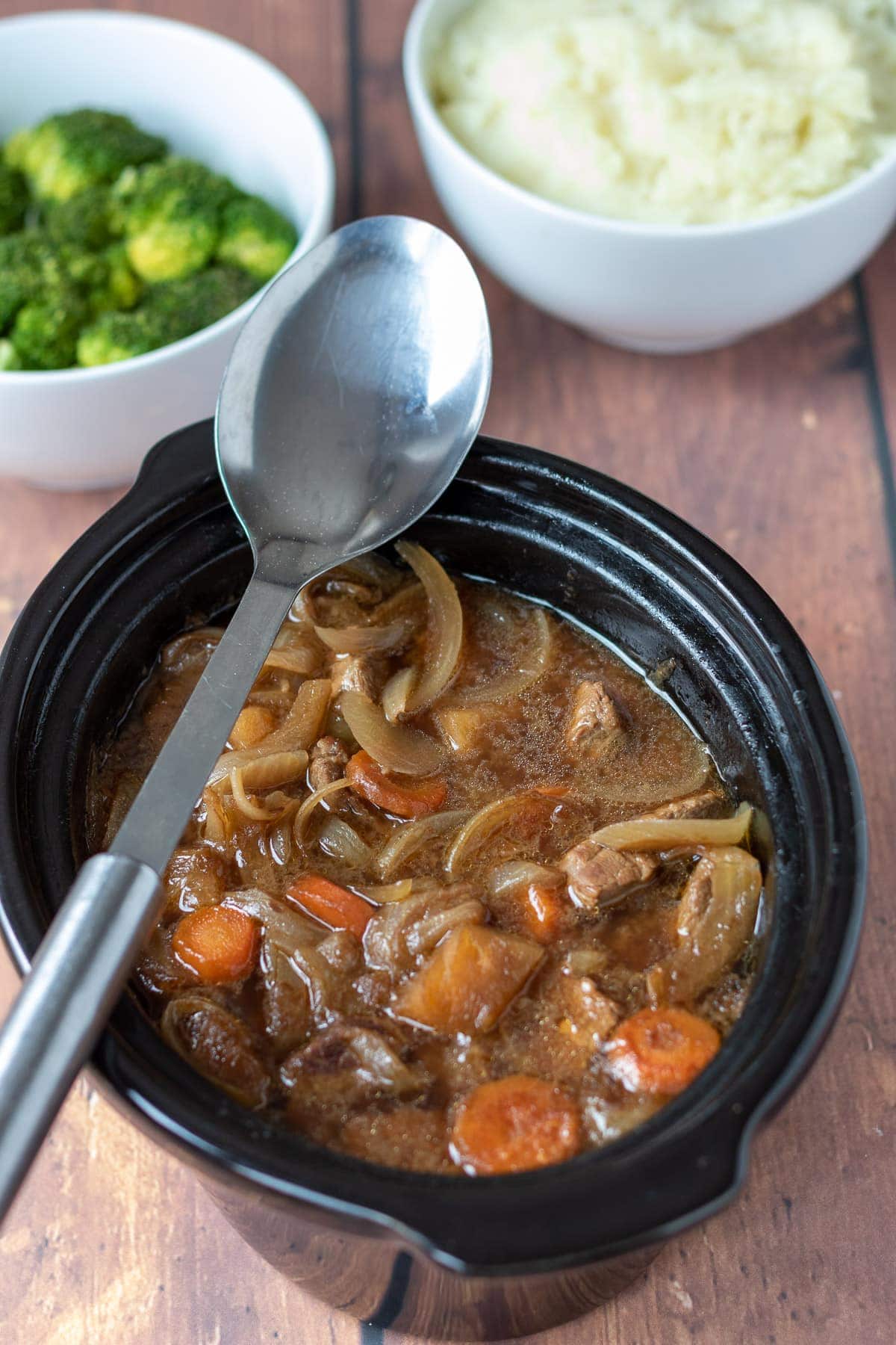 Slow cooker beef and ale stew with a serving spoon placed over the top. Side dishes of broccoli and mashed potatoes at the top.