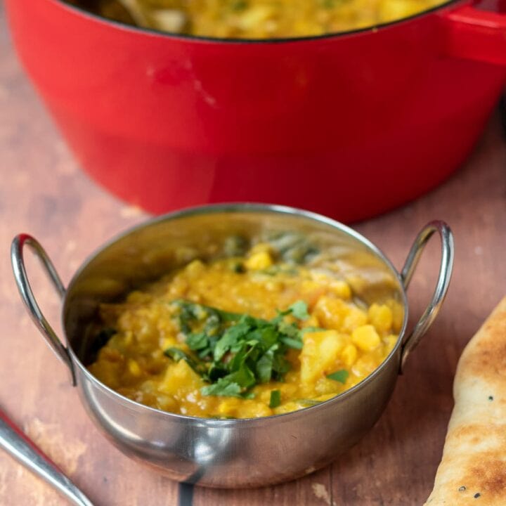 One pot vegetable dhal served in a balti dish with the rest in a casserole pot in the background. Spoon and Naan bread at the front.