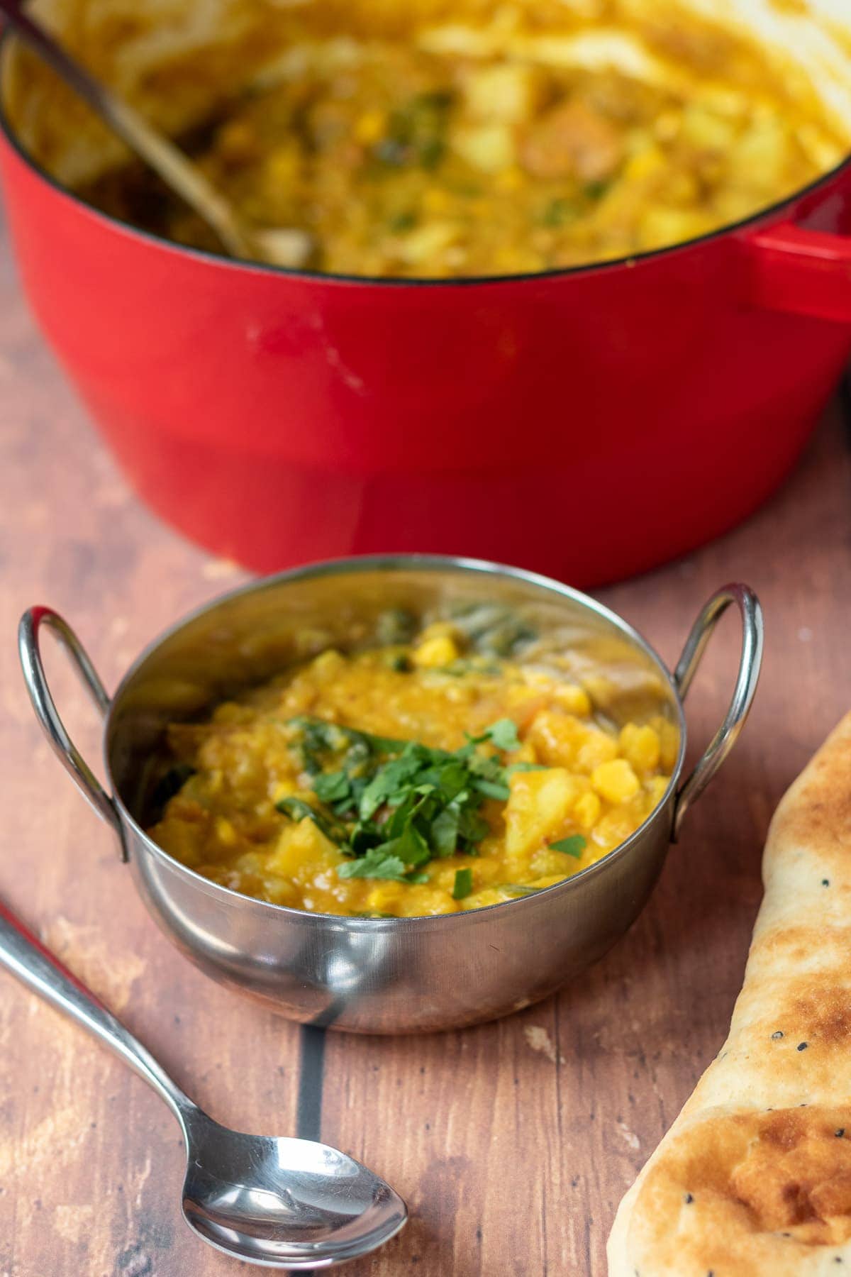 One pot vegetable dhal served in a balti dish with the rest in a casserole pot in the background. Spoon and Naan bread at the front.