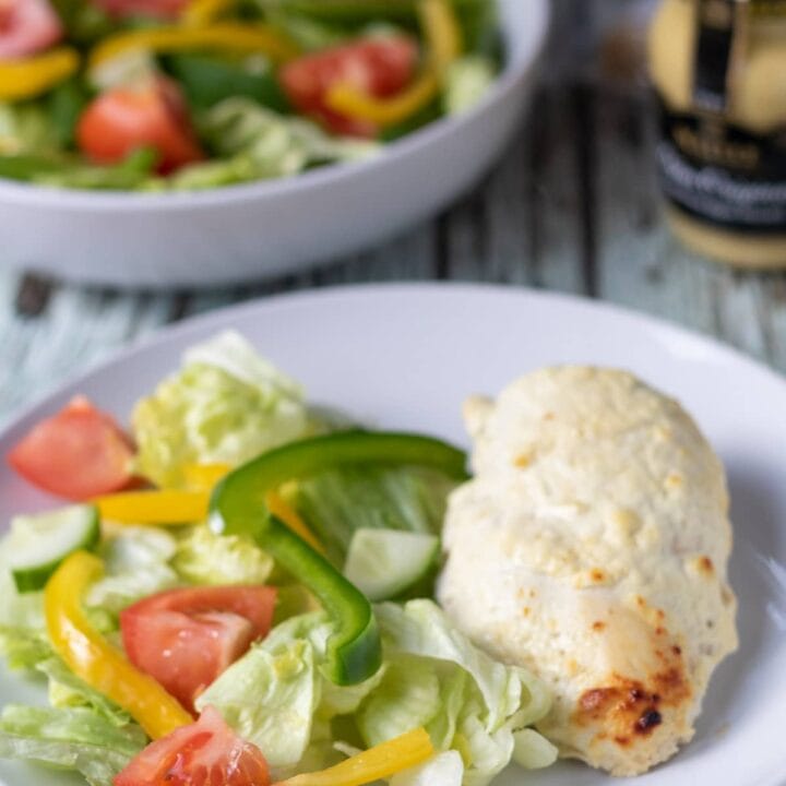 Oven baked mustard yogurt chicken served on a plate with salad. Jar of mustard and bowl of salad in the background.