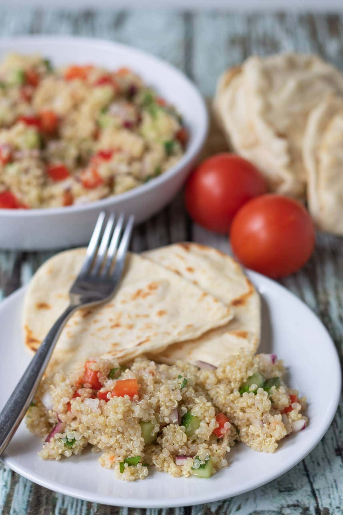 A plate with a serving of quick quinoa salad and flatbreads on as accompaniment. The rest of the salad in a bowl in the background with two tomatoes and flatbreads beside.