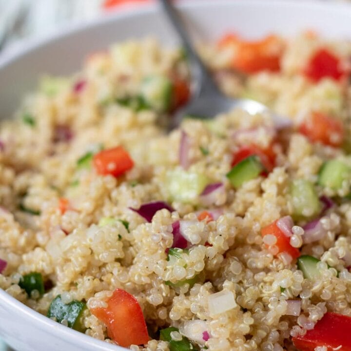 A white bowl of quick quinoa salad with a serving spoon in. Two tomatoes and a lemon in the background.