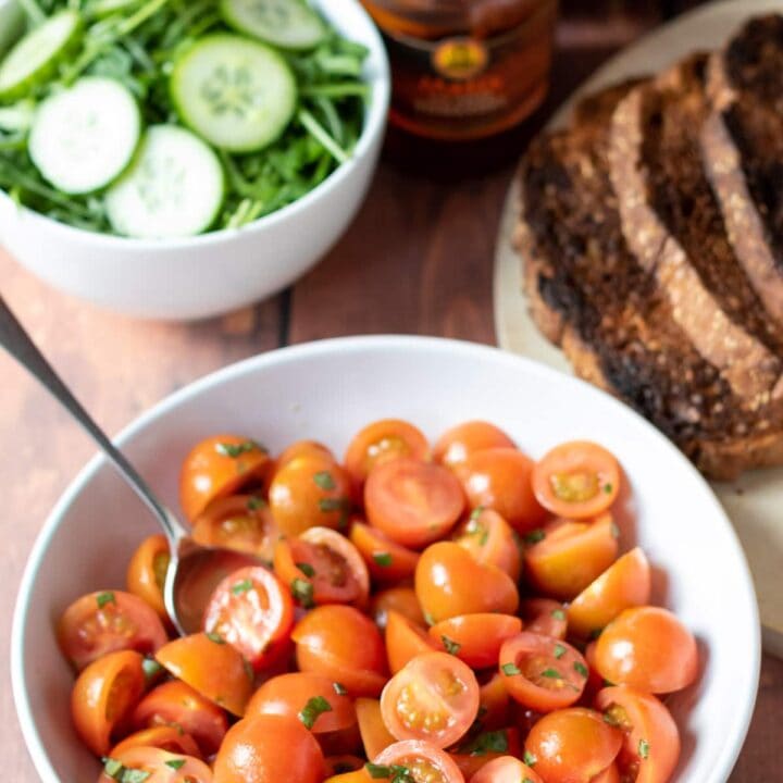 A bowl of cherry tomato salad with a bowl of cucumber, sliced bread and cold meat platter alongside.