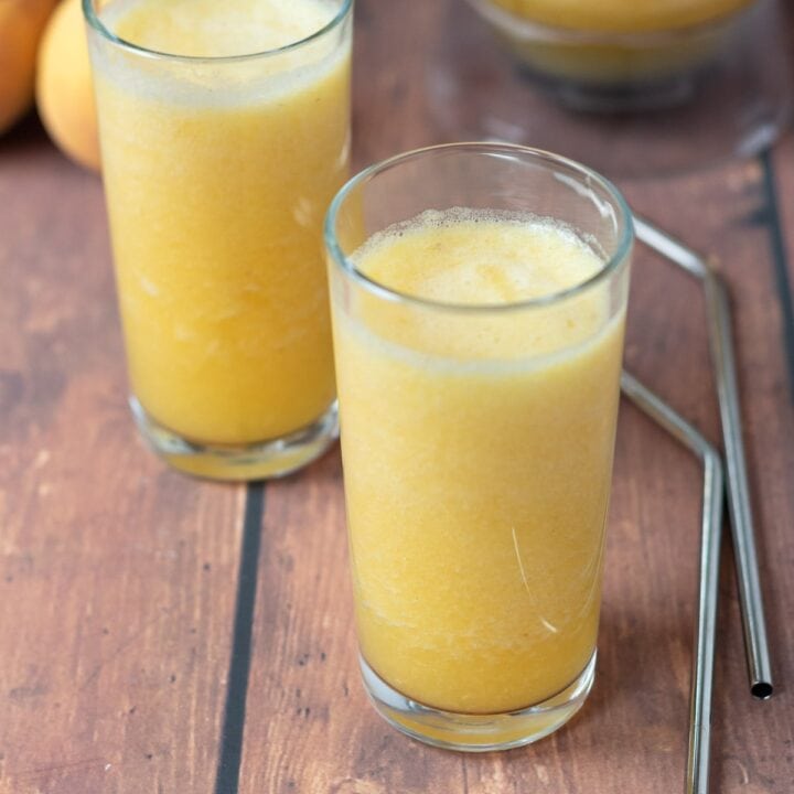 Two glasses of tropical pineapple smoothie diagonally across from each other. Straws to the side with the rest of the smoothie in a blender jug in the background with peaches beside.