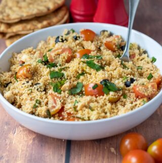 A serving bowl of couscous chicken salad with a spoon in. Cherry tomatoes to the front and salt and pepper shakers and flat bread behind.
