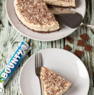 Birds eye view of a slice of no bake bounty coconut cheesecake on a plate with a fork to the side. Rest of the cheesecake at top of picture with a cake slice.