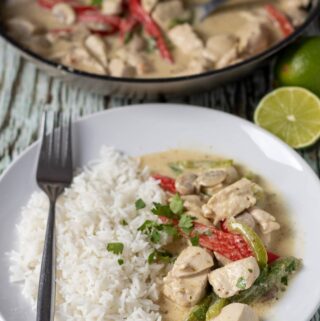 A plate of easy thai green chicken curry served with basmati rice and a fork to the side. Pan of cooked curry in the background.