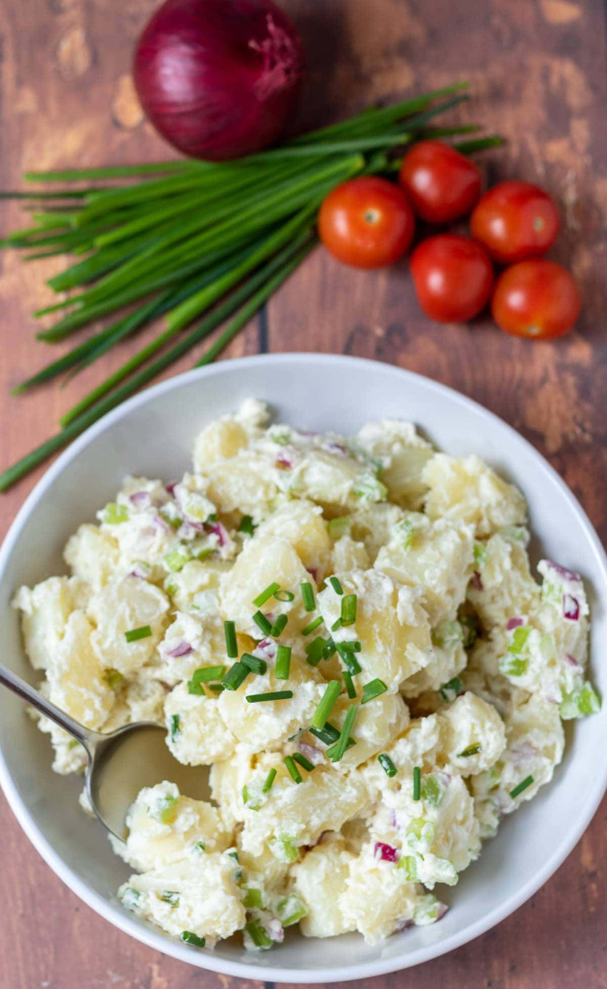 Birds eye view of a bowl of healthy potato salad at the bottom. Chives, cherry tomatoes and a red onion at the top.