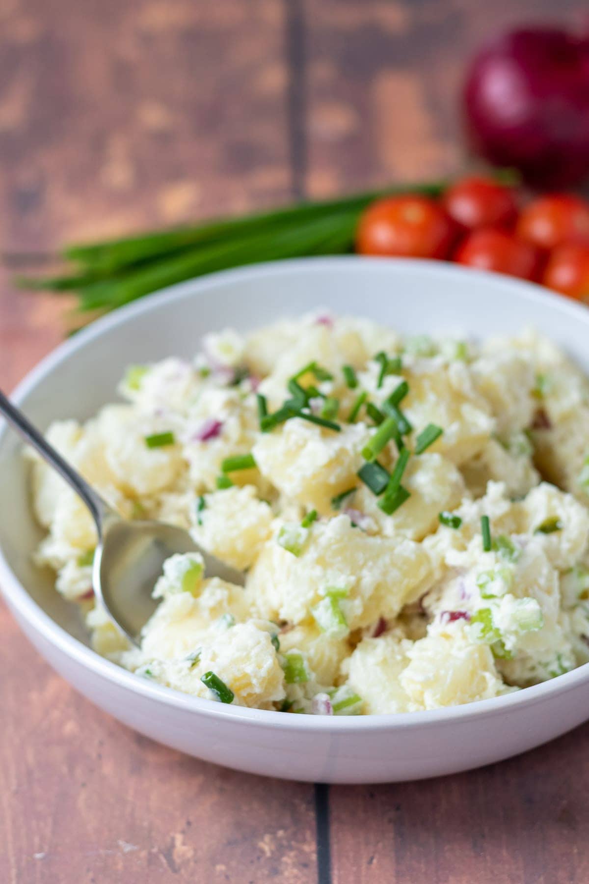 Potato salad in a bowl with a serving spoon to the side. Chives, cherry tomatoes and a red onion in the background as decoration.