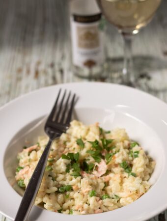 A plate of creamy salmon risotto with peas garnished with chopped parlsey and a fork to the side. A glass of wine and bottle in the background.