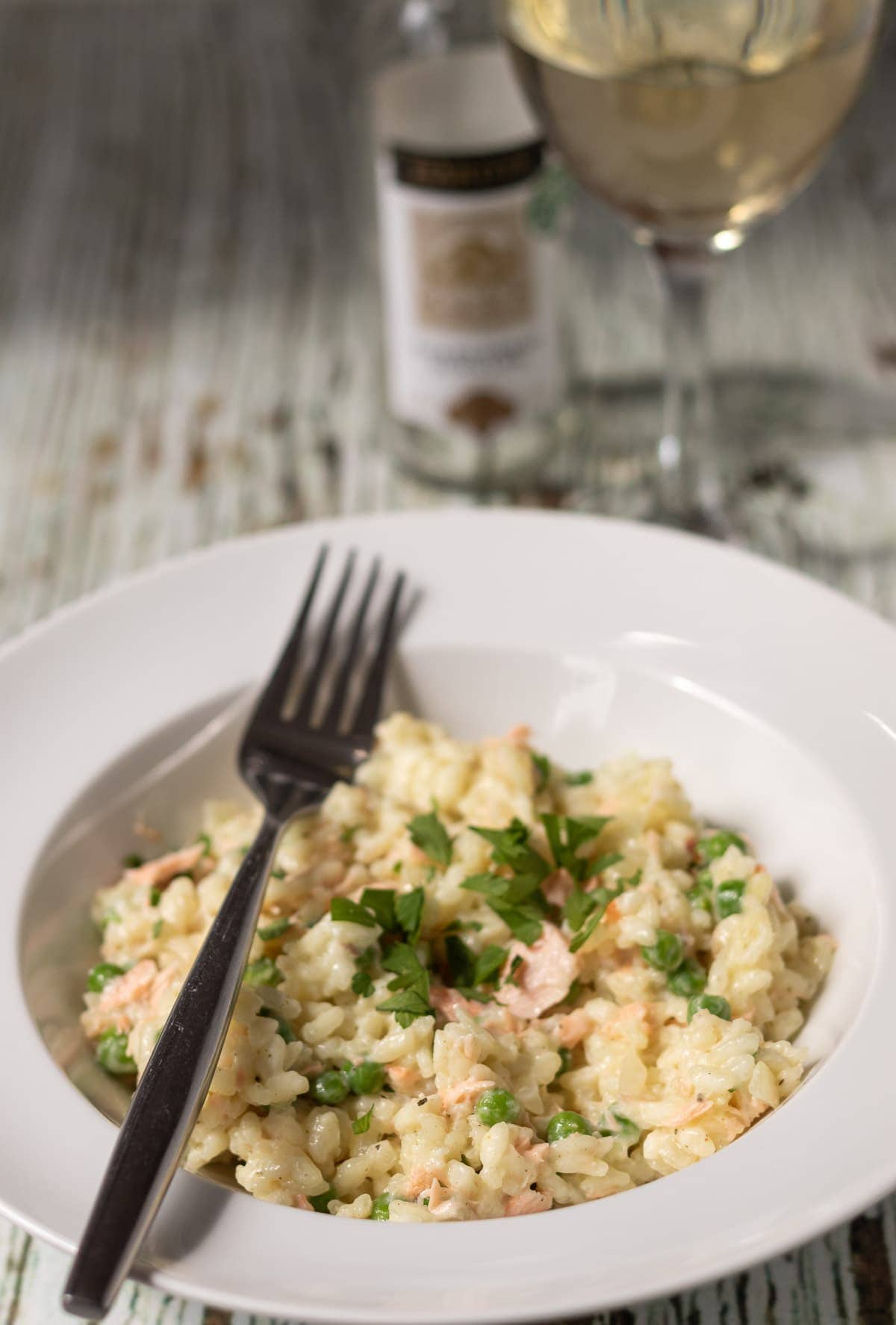 A plate of creamy salmon risotto with peas garnished with chopped parlsey and a fork to the side. A glass of wine and bottle in the background.