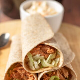 Three rolled slow cooker shredded BBQ chicken wraps stacked with a ramekin of coleslaw in the background.
