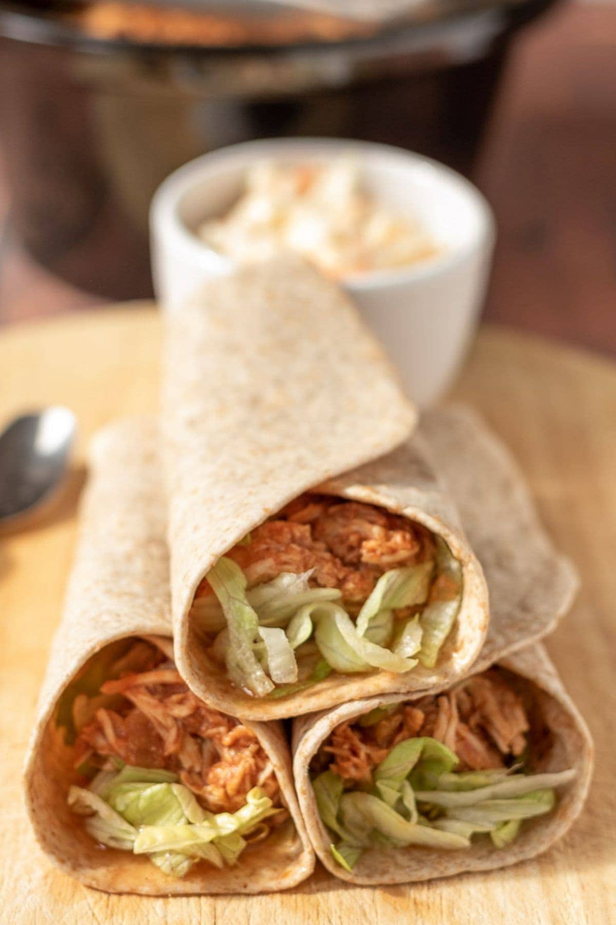 Three rolled slow cooker shredded BBQ chicken wraps stacked with a ramekin of coleslaw in the background.