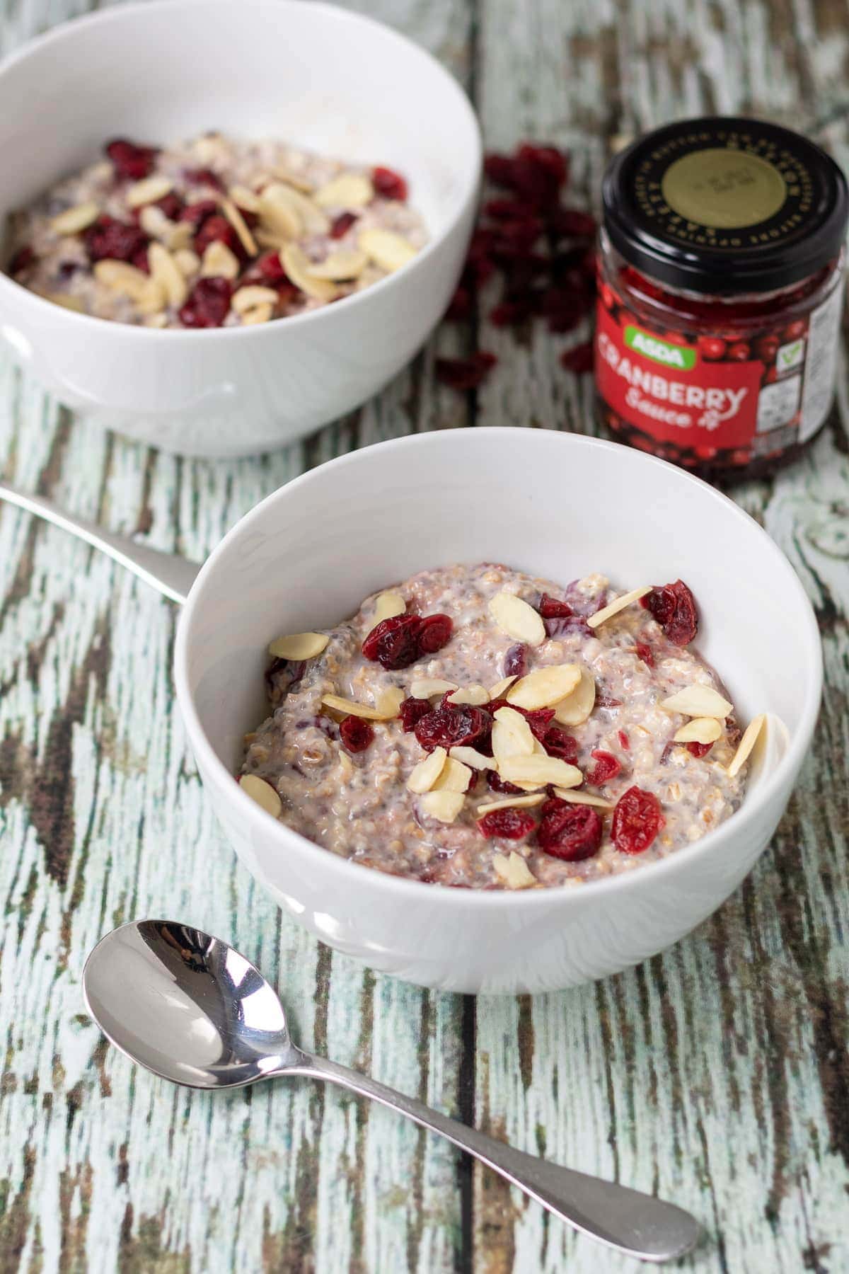 Two bowls of cranberry overnight oats with spoons between. A jar of cranberry sauce at top right.