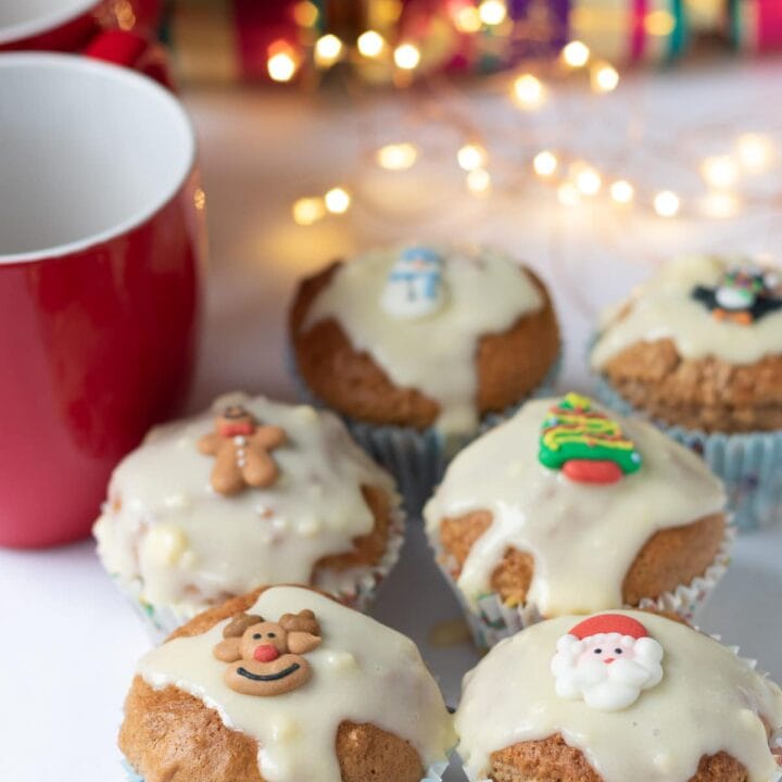 Six gingerbread muffins with cream cheese topped with christmas decorations. Two red mugs and Christmas crackers and festive lights in the background.