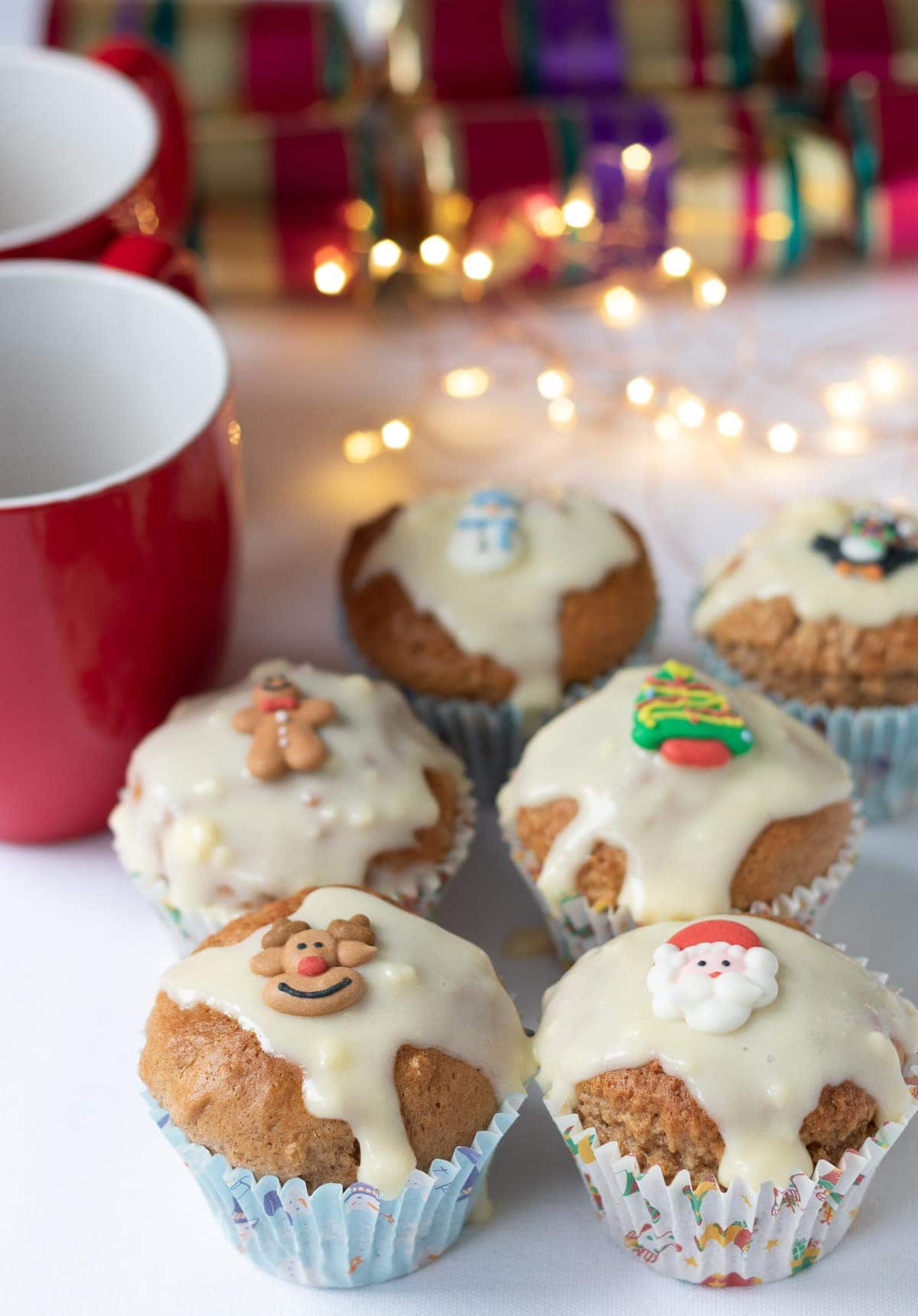 Six gingerbread muffins with cream cheese topped with christmas decorations. Two red mugs and Christmas crackers and festive lights in the background.