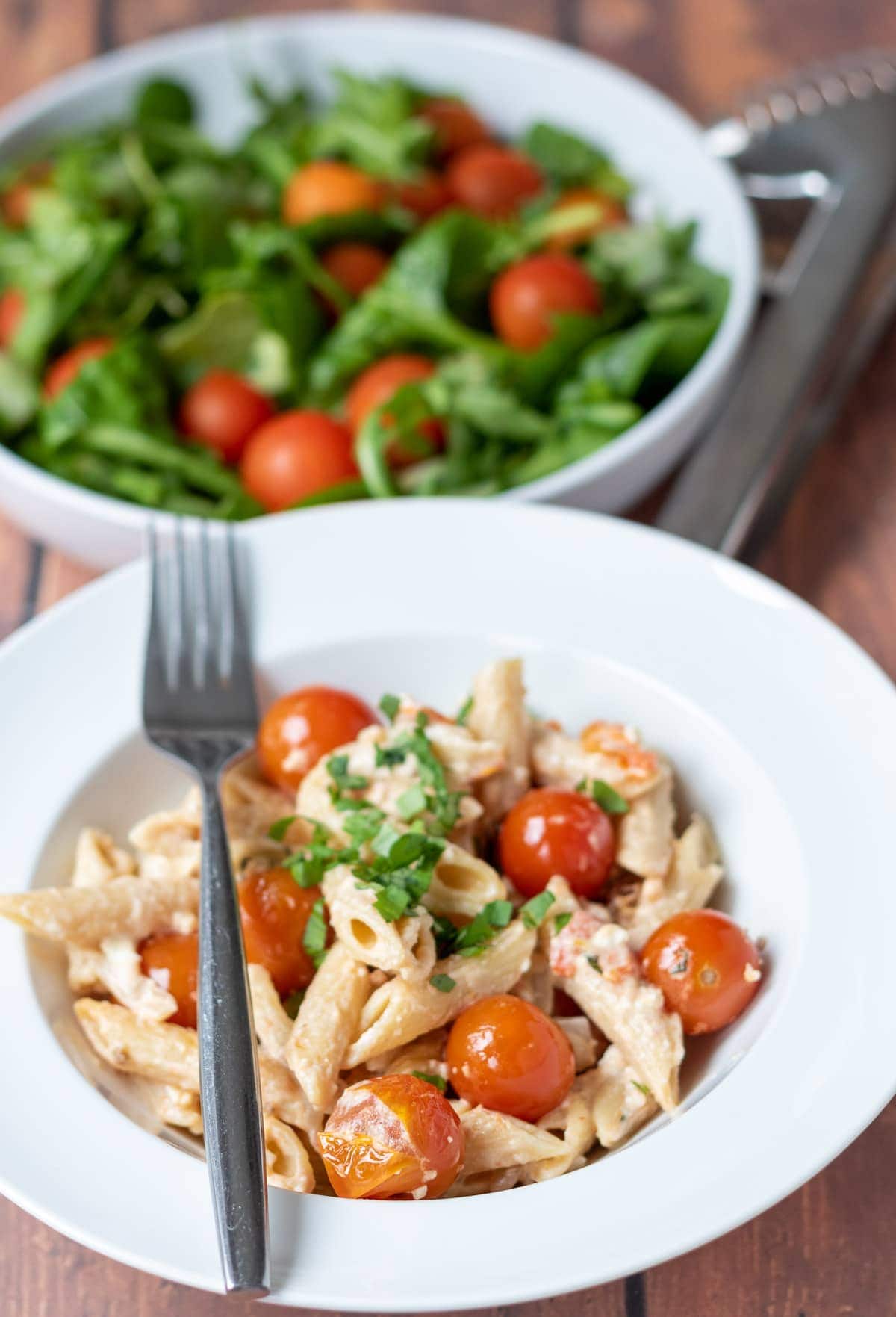 A pasta bowl with baked feta pasta in and a fork to the side. Side salad at the top of image with salad serving tongs.