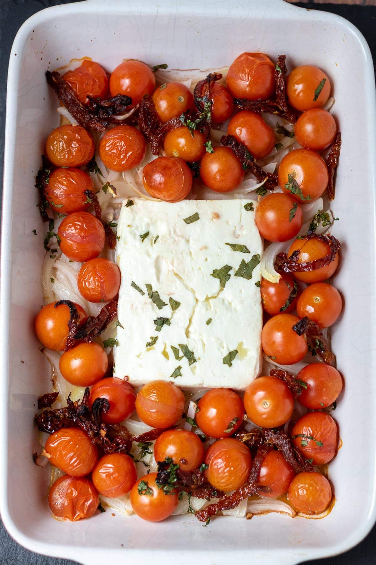Step 2 of images showing how to make healthy baked feta pasta. A stoneware dish with a block of feta cheeese in and cherry and sundried tomatoes and olive oil and red wine vinegar around.