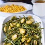 Roasted brussels sprouts and green beans served on a large platter dish with a dish of sweetcorn, jug of gravy and roast chicken in the background.
