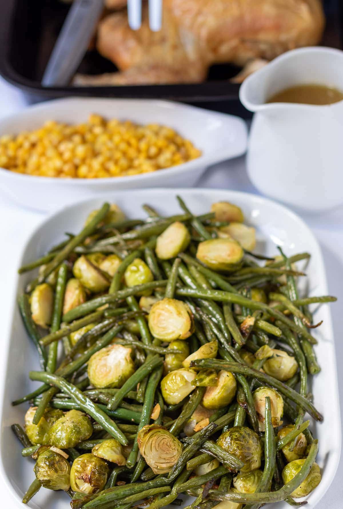 Roasted brussels sprouts and green beans served on a large platter dish with a dish of sweetcorn, jug of gravy and roast chicken in the background.