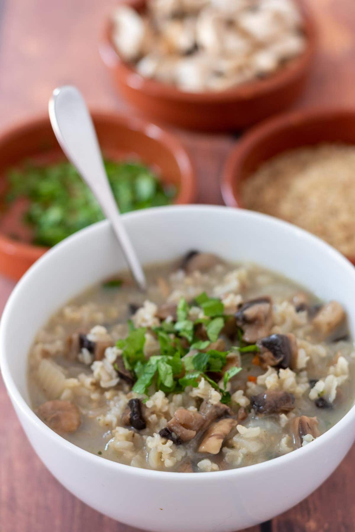 A bowl of mushroom and rice soup with a soup spoon in. Small dishes of chopped parsley, brown rice and chopped mushrooms in the background.