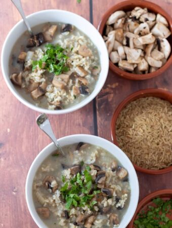 Birds eye view of two bowls of healthy mushroom and rice soup with soup spoon in. Small dishes of chopped parsley, brown rice and sliced mushrooms beside.