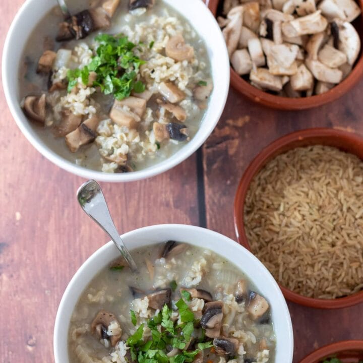 Birds eye view of two bowls of healthy mushroom and rice soup with soup spoon in. Small dishes of chopped parsley, brown rice and sliced mushrooms beside.