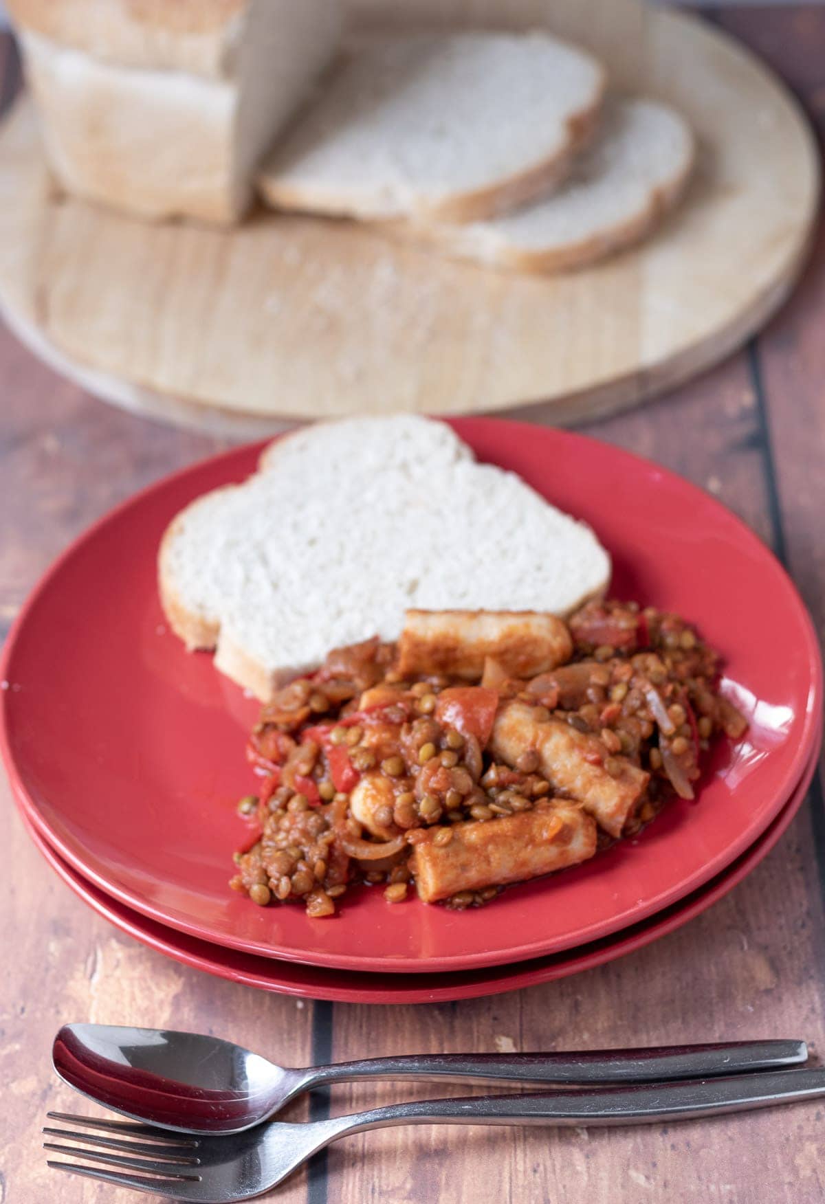 Sausage and lentil casserole served on a plate with a slice of bread. Spoon to the front and a loaf of bread cut on a board in the background.
