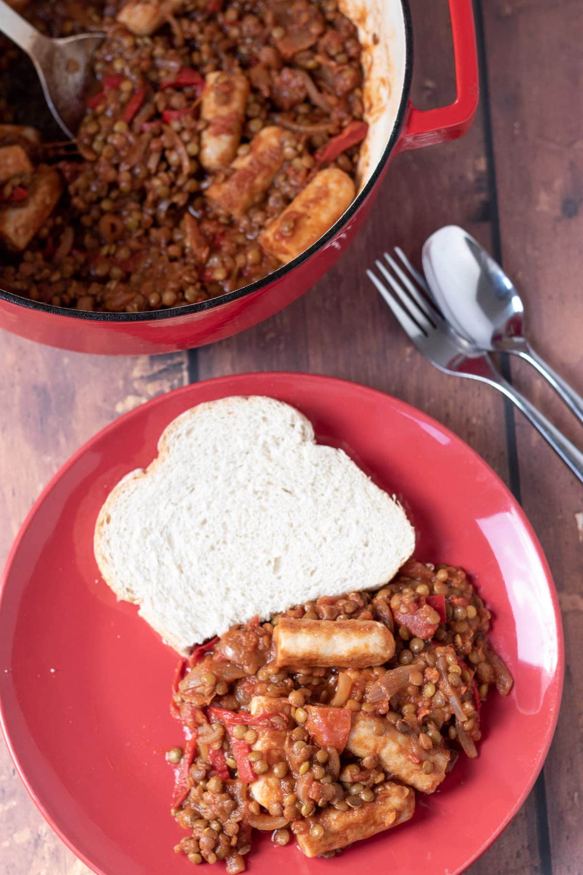 Birds eye view of a plate of sausage lentil casserole with a slice of bread. Rest of the casserole in a casserole pot and a knife and fork to the side.