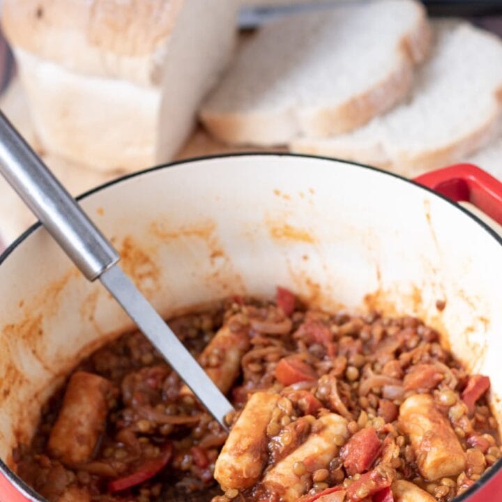 One pot sausage and lentil stew in a red casserole pot with a spoon in. A loaf of bread with slices cut off in the background.