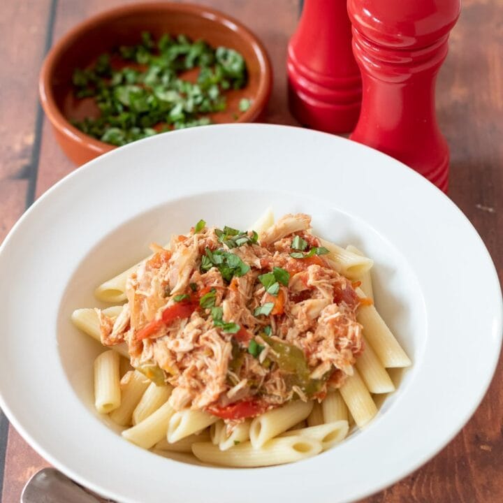 Slow cooker Italian chicken served over a bed of penne pasta in a white pasta bowl. A spoon to the front and a dish of chopped basil and salt and pepper shakers to the rear.
