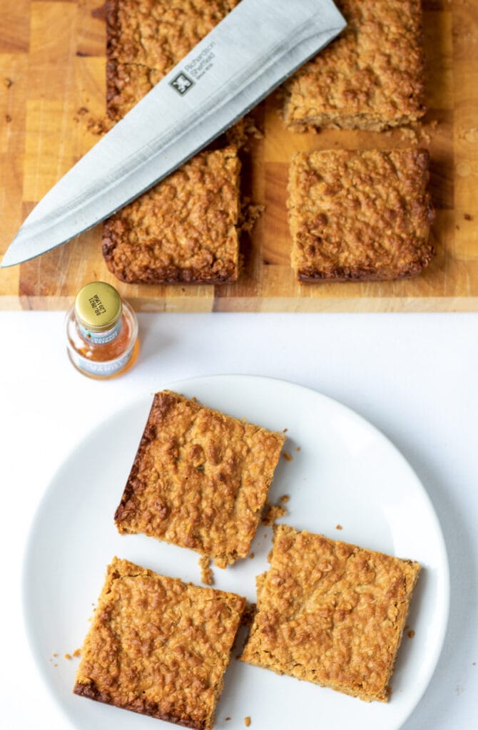 Birds eye view of three easy healthier flapjacks on a plate with the rest of the flapjacks on a chopping board with a knife at top.