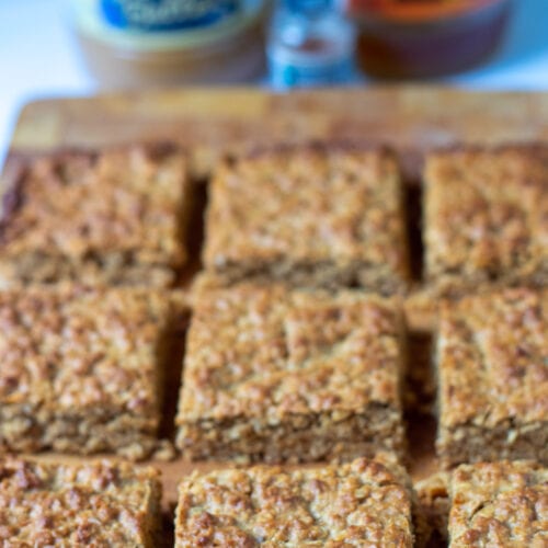 Easy healthier flapjacks recipe baked and cut into nine squares on a board. Jar of peanut butter and jar of honey in the background.