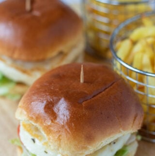 Two spicy red pesto halloumi burgers on a bread board with two basked of fries in the background.