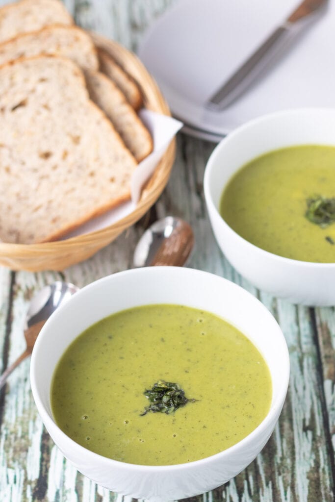 Two bowls of pea and basil soup with slices of bread in a basket and plates in the background.