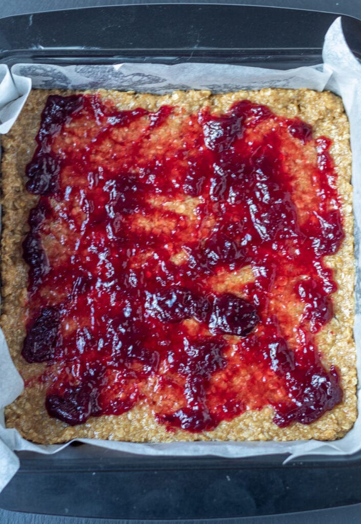 Cherry jam spread over half of the oat mixture in the square baking tin.