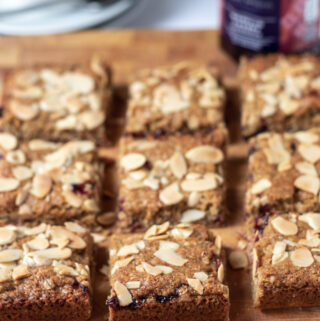 Cherry bakewell flapjacks cut into 9 squares on a chopping board with a teaspoon of cherry jam in front. Cup of tea and jar of jam in the background.