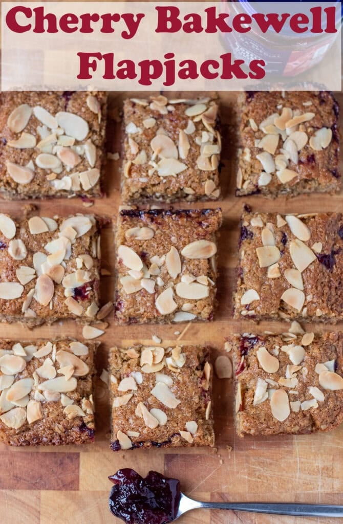 Overhead view of 9 cherry bakewell flapjack squares on a wooden chopping board. Teaspoon with jam in at the bottom. Pin title text overlay at top.